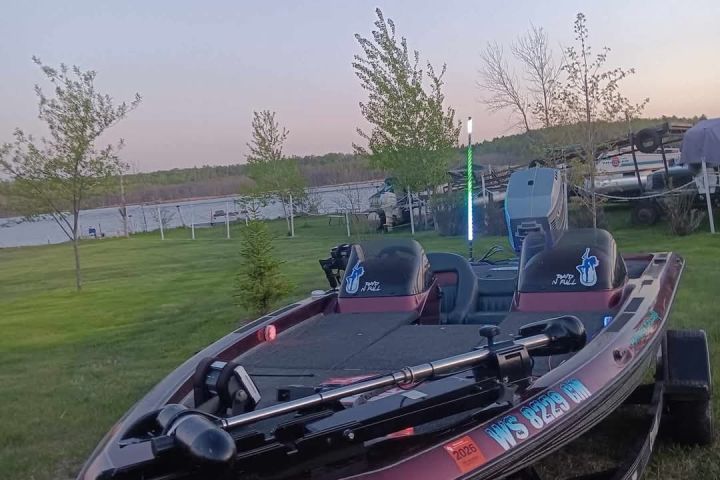 A maroon boat on a trailer near a lake with trees and a clear sky.