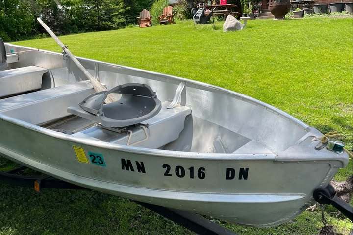 Small aluminum boat on grass with a house and trees in the background on a sunny day.