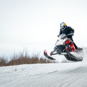 Person on a snowmobile jumping over a snow-covered hill with bare trees in the background.