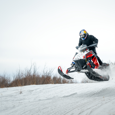 Person on a snowmobile jumping over a snow-covered hill with bare trees in the background.