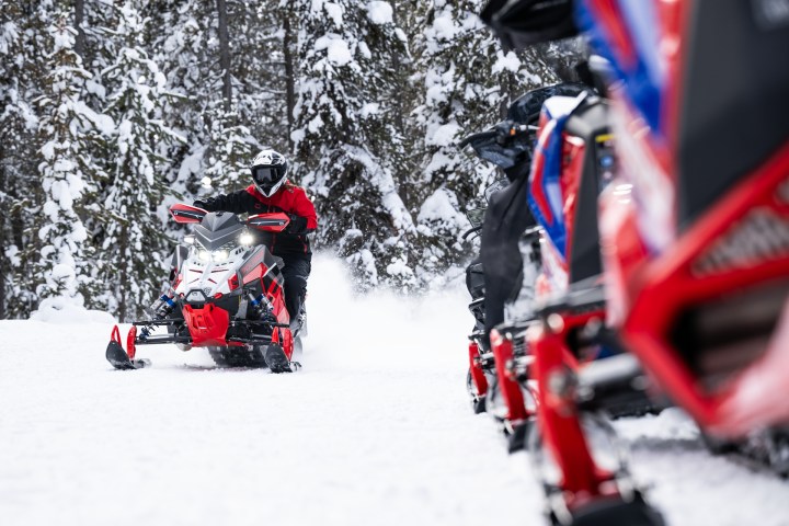 Rider on a red snowmobile in snowy forest, approaching camera.
