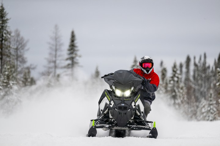 Person in red jacket rides a snowmobile on a snowy trail with trees in the background.