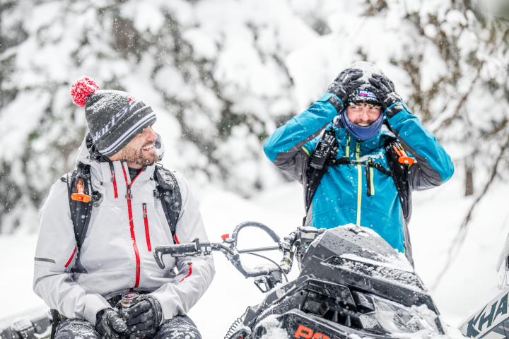 Two men in winter gear with snowmobiles, one adjusting helmet, in snowy forest setting.