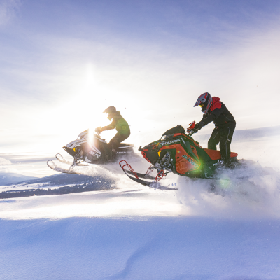 Two people riding snowmobiles across snow-covered landscape against a bright sun.