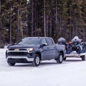 Truck towing snowmobiles on a snowy road in a forested area.