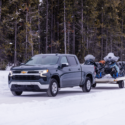 Truck towing snowmobiles on a snowy road in a forested area.