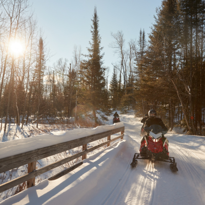 Two people riding snowmobiles on a snowy trail through a forest under a bright sun.