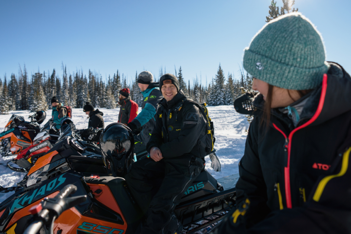 Group of people in winter gear with snowmobiles on snowy terrain under a clear blue sky.
