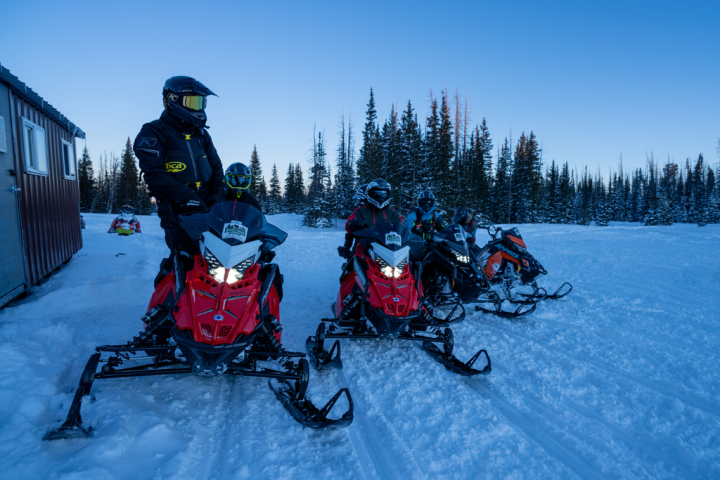 Four snowmobile riders in winter gear parked on snowy terrain near a cabin and pine trees.