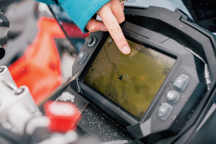 Hand pointing at GPS screen on snowmobile with snowy background.