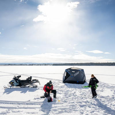 Two people ice fishing with a tent and snowmobile on a snowy frozen lake under a clear sky.