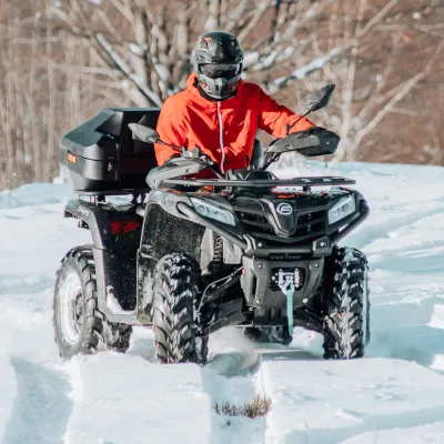 Person in red jacket riding an ATV in snowy landscape with bare trees.
