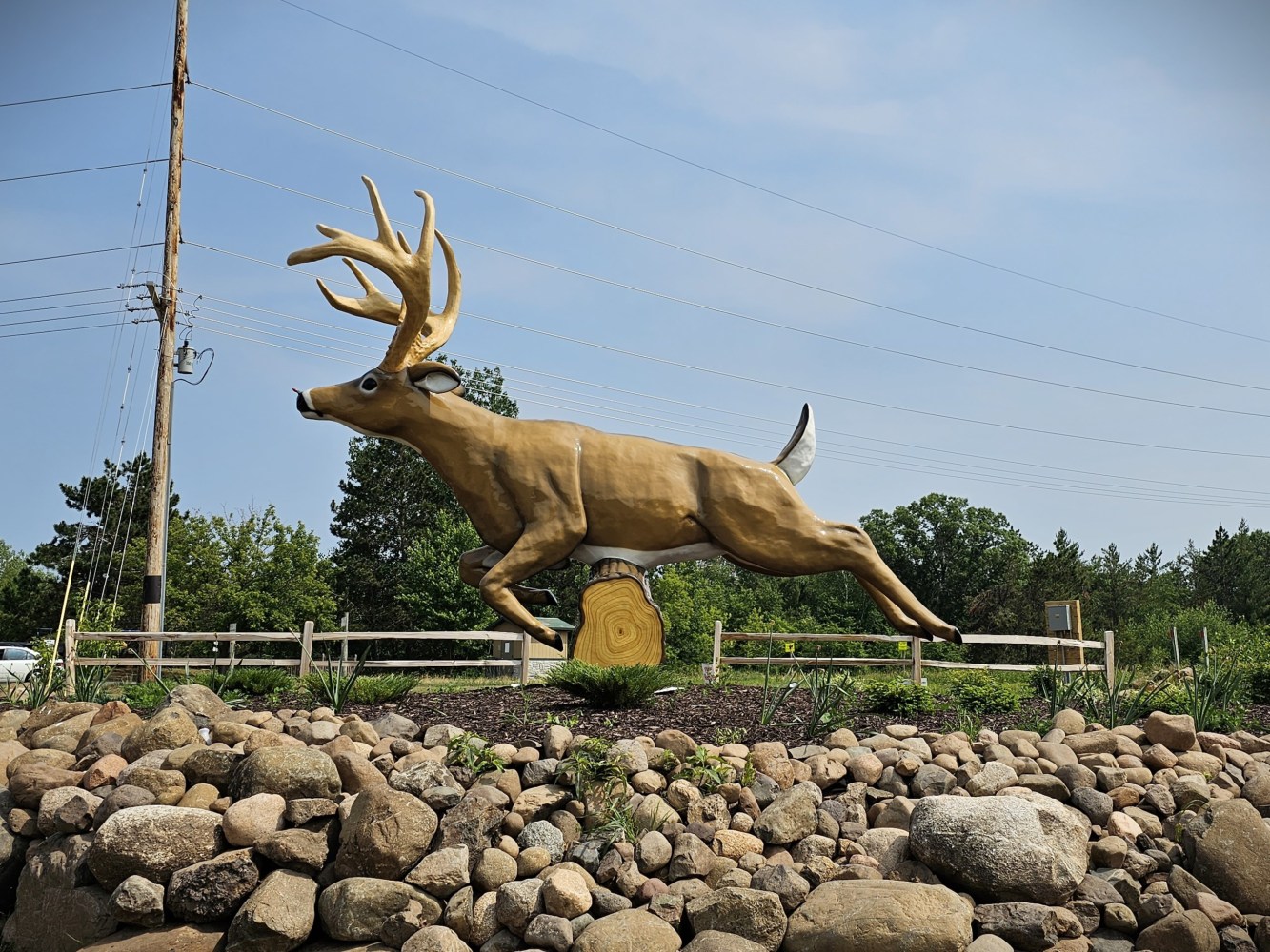 Large jumping deer sculpture on rock-lined base near wood fence and trees.
