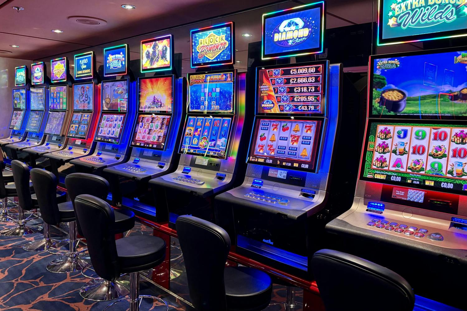 Row of colorful slot machines in a casino with black chairs.